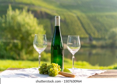 Famous German Quality White Wine Riesling, Produced In Mosel Wine Regio From White Grapes Growing On Slopes Of Hills In Mosel River Valley In Germany, Bottle And Glasses Served Outside In Mosel Valley