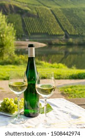 Famous German Quality White Wine Riesling, Produced In Mosel Wine Regio From White Grapes Growing On Slopes Of Hills In Mosel River Valley In Germany, Bottle And Glasses Served Outside In Mosel Valley