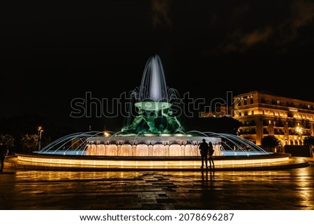 Famous illuminated Tritons Fountain at night,three bronze mythological Tritons sculptures holding up huge basin,modern landmark of Valletta.Long exposure photo,blurred people in motion.Night city