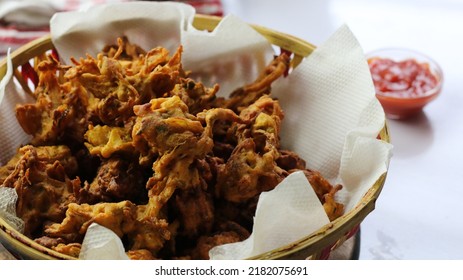 Famous Indian Monsoon Snack Known As Kanda Bhajji Or Pyaaz Pakode Is Served In Bamboo Basket With Tomato Sauce In The Background. (Indian Street Food Concept)