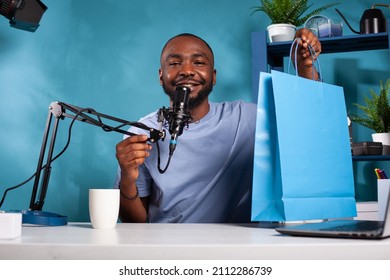 Famous influencer holding microphone recording podcast presenting giveaway blue paper gift bag in vlogging studio with professional setup. Content creator making video for sponsor giveaway.