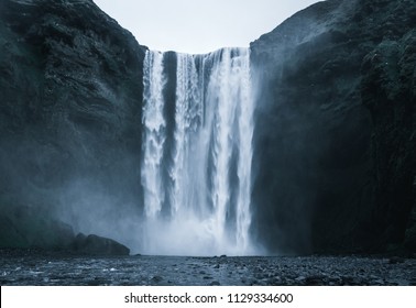 Famous Skogafoss Waterfall in Iceland from the front with rough looking water falling off the hill in a dark look with black stones in the foreground and no people in front during night