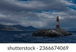 The famous southernmost old lighthouse Les Eclaireurs  in the Beagle Channel. A stone tower with red and white stripes stands on a rocky island against the sky and clouds. Mountains in the distance