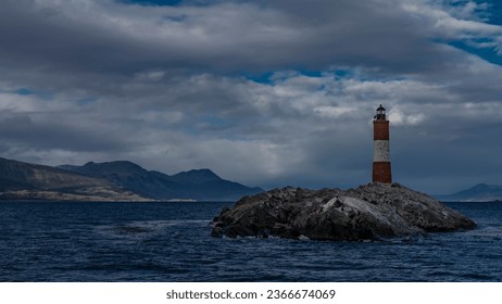 The famous southernmost old lighthouse Les Eclaireurs  in the Beagle Channel. A stone tower with red and white stripes stands on a rocky island against the sky and clouds. Mountains in the distance - Powered by Shutterstock