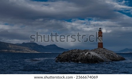 The famous southernmost old lighthouse Les Eclaireurs  in the Beagle Channel. A stone tower with red and white stripes stands on a rocky island against the sky and clouds. Mountains in the distance