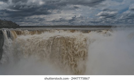 The famous waterfall is the Devil's Throat. Streams of bubbling foaming water collapse into the abyss. Splashes, fog. Clouds in the blue sky. Green vegetation on the river bank. Iguazu Falls.Argentina