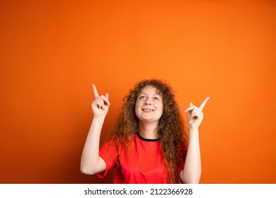 Fan Football Woman In Red T-shirt Team Uniform Looking Up And Keeping Two Index Fingers Up Posing On Orange Background.