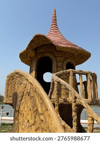 A fanciful playground structure designed to look like a rustic, fairy-tale tower. The sculpture features a textured, tree-bark finish with a spiral, pointed roof and a small balcony. It is set against a clear blue sky, emphasizing its whimsical and imaginative architecture.

