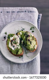 Fancy Avocado Toast With Micro Green And Poached Egg. Egg Yolk Pouring On Plate. Minimalist Food Styling. Grey Background. Avocado Slices On Sourdough Bread. Micro Greens. Healthy Breakfast.