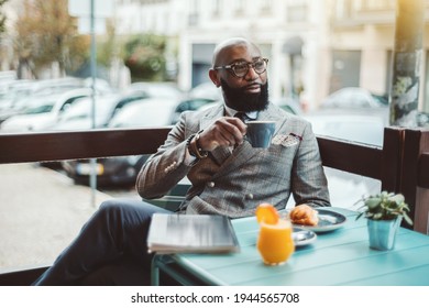 A fancy bearded hairless senior entrepreneur in glasses and a stylish custom-made costume, is sitting in an outdoor cafe in the morning and drinking a delicious coffee and pensively looking aside
