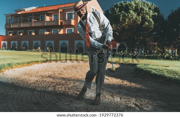 A fancy dapper mature black guy in an elegant outfit\
with trousers with suspenders, hat, tie, with a cigar in his mouth,\
is hitting the ball from a sand bunker zone of a golf field using\
an iron club