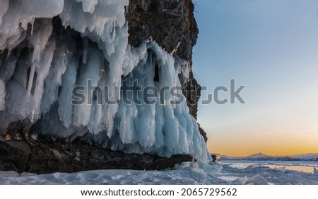 Fancy icicles hang from the base of the granite rock. The evening sky is highlighted in orange. The glare of the sun on a frozen lake. Baikal.