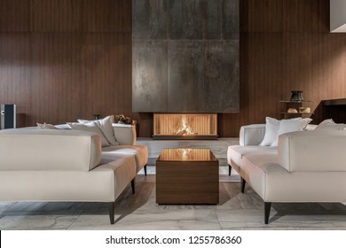 Fancy modern room with a burning fireplace, wooden wall and a tiled floor. There is a glossy table with sofas with pillows, glass stand with vases, black bag with firewood, speaker. Horizontal. - Shutterstock ID 1255786360
