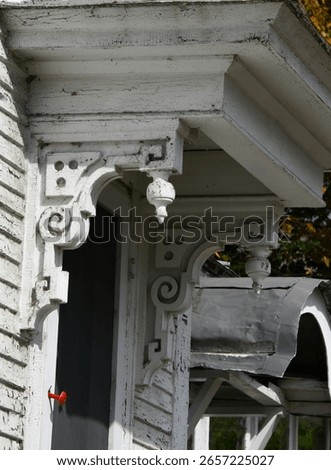 Fancy scroll work embellishes the eaves of olf wooden homes' porch.  Paint is cracked, peeling and dirty with cob webs.  Red door handle is barely hanging by a nail.