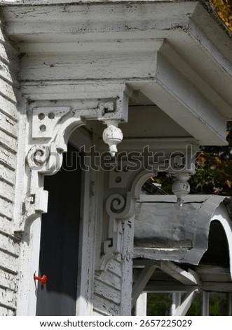 Fancy scroll work embellishes the eaves of olf wooden homes' porch.  Paint is cracked, peeling and dirty with cob webs.  Red door handle is barely hanging by a nail.