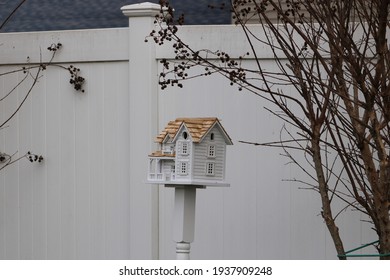 A fancy, wooden colonial birdhouse on a stake in a suburban backyard. The bird house is white with unpainted cedar shingles for a roof. The birdhouse is in front of a white PVC fence and under a bush.