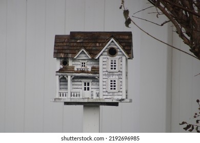 A Fancy, Wooden Colonial Birdhouse On A Stake In A Suburban Backyard. The Bird House Is White With Unpainted Cedar Shingles For A Roof. The Birdhouse Is In Front Of A White PVC Fence And Under A Bush.