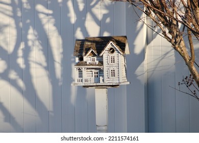 A Fancy, Wooden Colonial Birdhouse On A Stake In A Suburban Backyard. The Bird House Is White With Unpainted Cedar Shingles For A Roof. The Birdhouse Is In Front Of A White PVC Fence And Under A Bush.