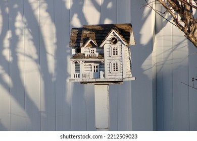 A Fancy, Wooden Colonial Birdhouse On A Stake In A Suburban Backyard. The Bird House Is White With Unpainted Cedar Shingles For A Roof. The Birdhouse Is In Front Of A White PVC Fence And Under A Bush.