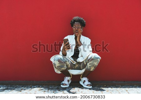 A fancy young African female is squatting near a red wall, making faces and texting via a smartphone; a funny black woman in sunglasses and with nail art is grimacing while making a video call