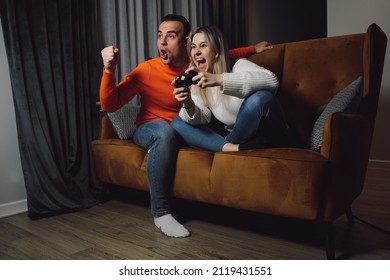 Fans Supporters Couple Dark-haired Smiling Enthusiastic Man And Blonde Woman Holding And Playing Sport Video Games Joystick Controller TV On Comfort Couch. Dark Gray Room Side View Indoor