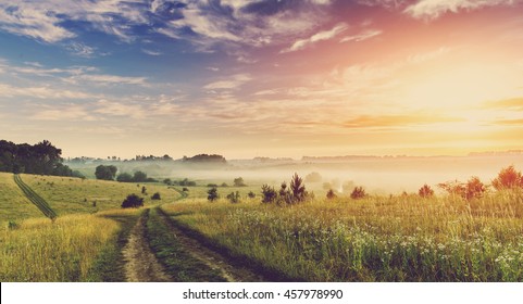 Fantastic foggy sunny day. ground road in the rural field with fresh grass in the sunlight. majestic misty sunrise with colorful clouds on the sky, Dramatic picturesque scene. retro style. 