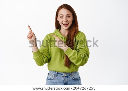 Fantastic promo ahead. Smiling redhead teen girl shows information, pointing left at advertisement and looking happy, standing over white background