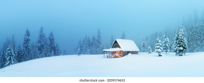 Fantastic Winter Landscape Panorama With Glowing Wooden Cabin In Snowy Forest. Cozy House In Carpathian Mountains. Christmas Holiday Concept