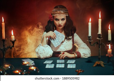 Fantasy beautiful girl in image of gypsy witch sits at table in dark gothic room. Art Red costume. Fortune teller magical woman reading future on tarot cards. Ritual candles burning, seance, smoke.