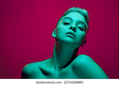 Fantasy. Beautiful young girl with short blonde hair posing with bare shoulders against pink studio background in green neon light. Cyberpunk style. Concept of futurism, digital world, robot, art