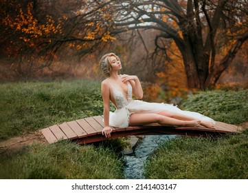 Fantasy Blonde Woman Nymph Fairy Princess Sitting On A Wooden Bridge. White Creative Design Fashion Dress Sexy Pose, Bare Legs. Autumn Forest Orange Color Trees Green Fresh Grass. Girl Enjoy Nature