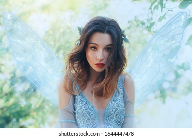 Fantasy closeup portrait attractive woman in image young fairy. carnival Costume blue dress transparent wings. face tender red makeup. Wavy Brunette hair Flutter fly in wind. Green nature backdrop