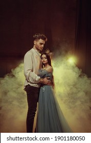 Fantasy couple hugging in dark room full white smoke. Image of lovers - king and queen. Medieval male prince in golden crown, vintage costume clothing. Girl Princess in long glamorous blue dress gown