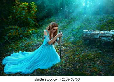 Fantasy elf woman princess warrior sits in forest on green grass holding weapon medieval sword in hands. Warlike queen redhaired girl in blue medieval dress. Trees, divine sun rays blessing, light fog