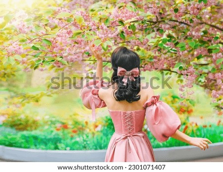 Fantasy girl princess hand touching flowers sakura tree spring nature green grass petals fall. Woman queen back rear view long pink dress puffed sleeves satin bow in hair vintage old style art photo.