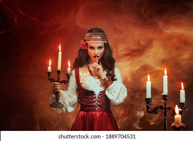 Fantasy Gypsy woman in red vintage dress. Girl witch fortune teller holds candlestick burning candles. Room in magical fire. Spiritual Art photo. finger touches lips gesture of secret mystery silence