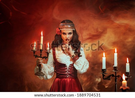 Fantasy Gypsy woman in red vintage dress. Girl witch fortune teller holds candlestick burning candles. Room in magical fire. Spiritual Art photo. finger touches lips gesture of secret mystery silence