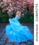 Fantasy happy woman princess in long fluffy blue dress, like Cinderella, circles in dance, fabric flies waves in motion. Fairy girl goddess blond hair. Spring blooming nature, sakura tree pink flowers