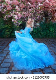 Fantasy happy woman princess in long fluffy blue dress, like Cinderella, circles in dance, fabric flies waves in motion. Fairy girl goddess blond hair. Spring blooming nature, sakura tree pink flowers