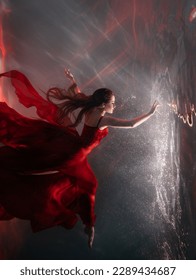 Fantasy mystery woman swims underwater touches magic light mirror, looks into reflection. Fairy tale Beauty Girl princess sleeping soars floating in dream dark water. Art ballerina dancing red dress.