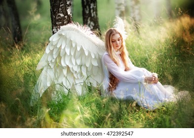Fantasy photo of beautiful angel in vintage dress sitting near tree.Amazing blond girl with long curly hair and white wings resting in sun light.Art work and fairy tale. Summer day.