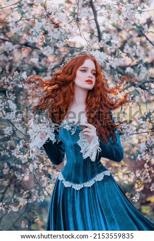 Fantasy portrait red-haired girl romantic princess stands in spring flowering garden. Blooming green tree flowers. Long hair red lips pale skin face woman queen medieval vintage creative design dress