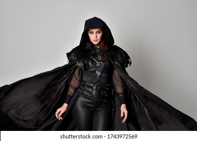 Fantasy Portrait Of A Woman With Red Hair Wearing Dark Leather Assassin Costume With Long Black Cloak. Close Up, 3/4 Pose  Isolated Against A Studio Background.