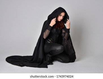 fantasy portrait of a woman with red hair wearing dark leather assassin costume with long black cloak. Full length kneeling pose  isolated against a studio background.