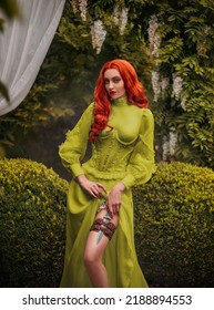 Fantasy Red-haired Dangerous Woman Medieval Assassin Queen With Dagger In Sheath On Leg Leather Belt. Green Vintage Old Style Dress. Girl Princess Warrior. Garden Summer Nature Trees. Red Long Hair