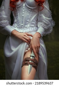 Fantasy Red-haired Dangerous Woman Medieval Assassin Queen With Dagger In Sheath On Leg Leather Belt. White Vintage Old Style Dress. Girl Princess Warrior. Green Summer Nature Trees. Hand Hold Knife