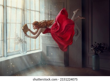 Fantasy redhead woman soars floats flies in air. Art photo levitation. Girl fairy princess reads magic book, divine light from window. Red midi dress, lon hair flutters in wind. Room classic interior - Shutterstock ID 2151224075