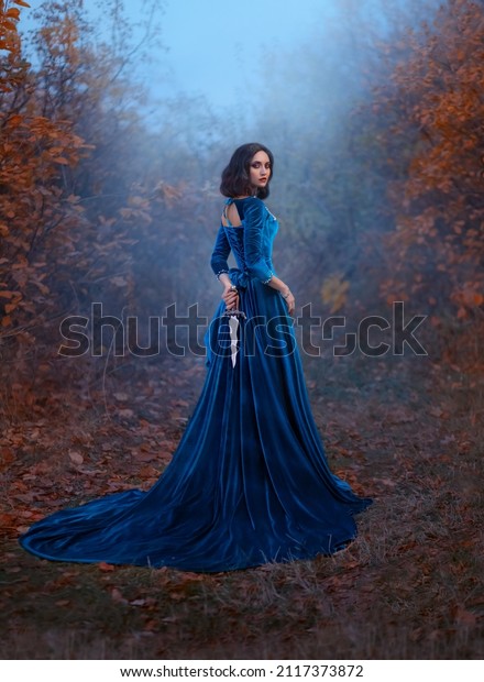 Fantasy warlike medieval woman queen holds metal\
vintage dagger blade weapon in hands hides behind back. Background\
forest fog night. Blue velvet luxurious royal dress. Girl princess\
warrior rear view