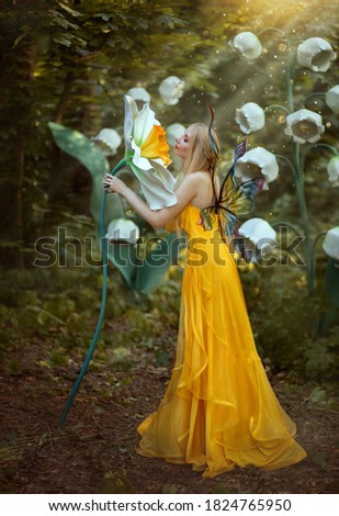 Fantasy woman blonde forest fairy. Fashion model in a long yellow dress with butterfly wings holds in hand and smell large flower narcissus. Scenery white lilies of the valley. Sun rays light magic.