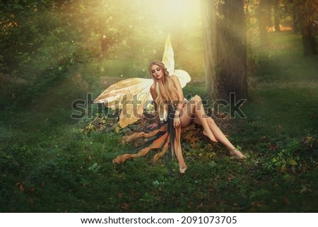 Fantasy woman fairy sits on green grass in fabolous summer forest. Girl goddess of nature. Golden pixie butterfly wings costume, green dress long legs. Fashion model posing in meadow, sun magic light